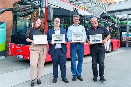 Vier Personen halten eine Urkunde und stehen vor einem Bus.