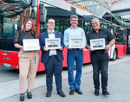 Vier Personen halten eine Urkunde und stehen vor einem Bus.